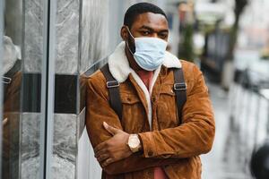 avvicinamento ritratto di un' elegante africano americano tipo sta solo nel il strada nel un' protettivo maschera su il suo viso. precauzionale le misure. foto