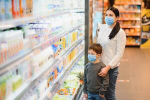 shopping con bambini durante virus scoppio. madre e bambino indossare chirurgico viso maschera acquisto frutta nel supermercato. mamma e poco ragazzo acquistare fresco verdura nel drogheria negozio. famiglia nel negozio foto