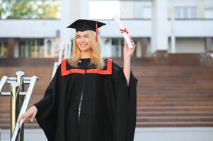 contento carino caucasico grad ragazza è sorridente. lei è nel un' nero mortaio asse, con rosso nappa, nel abito, con simpatico Marrone Riccio capelli, diploma nel mano foto