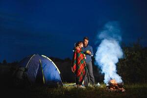 amorevole coppia escursionisti godendo ogni Altro, in piedi di fuoco di bivacco a notte sotto sera cielo vicino alberi e tenda. romantico campeggio vicino foresta nel il montagne foto