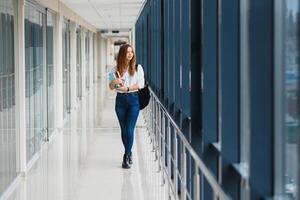 allegro brunetta alunno ragazza con nero zaino detiene libri nel moderno costruzione. femmina alunno in piedi con libri nel Università corridoio foto