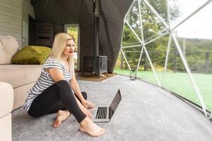 mezzo anziano donna usi un' il computer portatile riposo e la spesa tempo a glamping Casa su vacanze. vacanza cupola tenda. accogliente, campeggio, hygge, stile di vita concetto foto