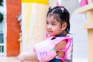 ragazza asiatica carina che gioca con l'acqua al resort. bambino che indossa tuta di sicurezza rosa. bambino che tiene tappetino di gomma gialla. bambini felici le bagnano la testa. tempo di relax. bambino di 4-5 anni. foto