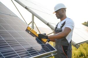 afroamericano lavoratore nel grigio tuta da lavoro e un' bianca difficile cappello lavori nel un' campo di solare pannelli. solare rinnovabile energia concetto. foto