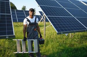 competente energia ingegnere nel grigio tuta da lavoro e bianca casco controllo solare pannelli mentre a piedi su campo. africano americano uomo trasporto appunti e contenitore con strumenti foto