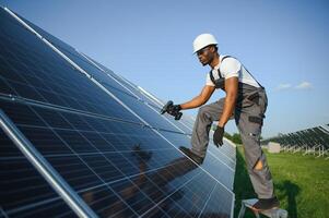 afroamericano lavoratore nel grigio tuta da lavoro e un' bianca difficile cappello lavori nel un' campo di solare pannelli. solare rinnovabile energia concetto. foto