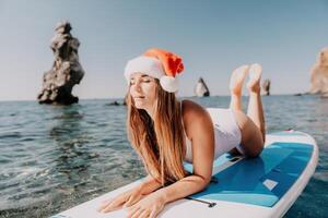 donna mare sup. vicino su ritratto di contento giovane caucasico donna con lungo capelli nel Santa cappello guardare a telecamera e sorridente. carino donna ritratto nel un' bianca bikini in posa su cenare tavola nel il mare foto