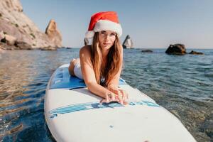 donna mare sup. vicino su ritratto di contento giovane caucasico donna con lungo capelli nel Santa cappello guardare a telecamera e sorridente. carino donna ritratto nel un' bianca bikini in posa su cenare tavola nel il mare foto