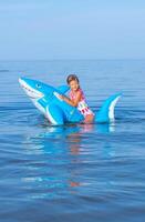 contento ragazza di europeo aspetto età di 7 nuoto su un gonfiabile grande squalo giocattolo nel il mare.famiglia estate vocazione concetto. verticale foto. foto