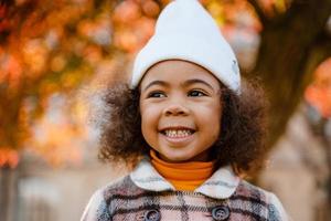 ragazza riccia nera che indossa un cappello bianco che sorride mentre cammina nel parco autunnale foto