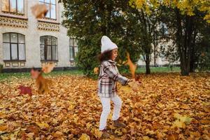 ragazza nera che indossa un cappotto che si diverte con le foglie cadute nel parco autunnale foto