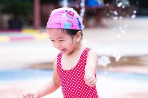 ragazza asiatica felice del bambino che gioca acqua. bambini che ridono. i bambini corrono nella fontana. il bambino indossava una cuffia rosa e un costume da bagno rosso con un motivo a rombi. bambino di 3 anni. foto