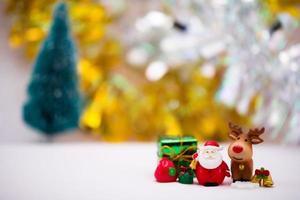 bambole in resina di babbo natale accanto a renne marroni. i lati sono decorati con scatole regalo, fiocchi di neve e campanelli d'oro. morbido pavimento bianco. bokeh debole su sfondo oro e argento e albero di natale foto