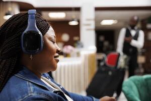 sorridente africano americano donna indossare senza fili cuffie riposo nel Hotel atrio. contento femmina turista ascoltando per musica mentre in attesa per registrare a ricorrere, in arrivo su vacanza foto