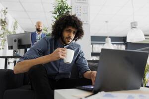 giovane arabo comapny dipendente potabile caffè e utilizzando aziendale il computer portatile nel attività commerciale ufficio. esecutivo manager Tenere tè boccale e soluzione opera compiti su computer nel coworking spazio foto