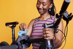 dettagliato Visualizza di nero donna l'esame danneggiato bicicletta telaio su riparazione In piedi contro giallo sfondo. africano americano signora fissaggio e fabbricazione necessario aggiustamenti su moderno bicicletta. foto