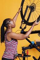 nel davanti di giallo sfondo, attivo giovane donna fa annuale Manutenzione e registrazione di bicicletta pneumatico. atletico africano americano signora protegge sua bicicletta ruota per all'aperto ricreativo Ciclismo. foto