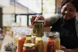 africano americano negoziante vende sottaceti a partire dal bicchiere barattoli nel sua zero rifiuto cibo mercato. sorridente nero donna indossare un grembiule mettendo plastica gratuito contenitore con conservato articolo su di legno contatore. foto