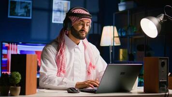 sorridente arabo uomo con senza fili cuffie a casa nel vivente camera, digitando messaggi su il computer portatile. contento musulmano persona ascoltando per musica, godendo rilassante tempo libero tempo parlando con amici su taccuino foto