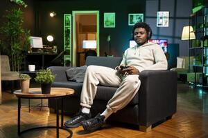 africano americano uomo nel debolmente illuminato appartamento giocando videogiochi, rilassante e avendo divertimento. gamer combattendo nemici nel multiplayer gioco su gioco consolle durante tempo libero tempo a casa foto