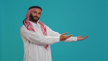 musulmano adulto nel tradizionale toga e ghutra presentazione lati con mani e in posa per studio sparo. Arabo gioioso uomo promozione qualcosa mentre mostrando marketing annuncio pubblicitario a parte. foto