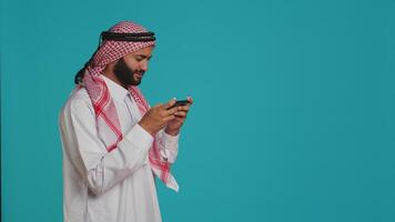 contento persona avendo divertimento con gioco su il suo smartphone dispositivo, indossare tradizionale Arabo abbigliamento. nel il studio, musulmano modello godendo e sport torneo con mobile Telefono videogiochi. foto