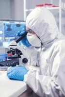 ricercatore nel moderno microbiologia laboratorio usi microscopio vestito nel ppe completo da uomo. virolog nel tuta durante coronavirus scoppio conduzione assistenza sanitaria scientifico analisi. foto