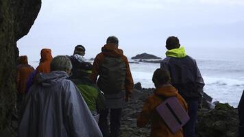 gruppo di turisti passeggiate su pietra costa. clip. turisti camminare su pericoloso pietra riva nel nuvoloso tempo atmosferico. gruppo di turisti su costa con nero rocce di settentrionale paesaggio foto