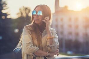 giovane elegante bella donna, mani Tenere un' Telefono. tramonto sfondo, soleggiato giornata, bene tempo atmosferico, occhiali da sole, freddo Accessori. foto