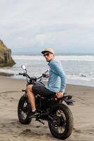 uomo nel berretto equitazione motociclo su spiaggia. moto attraversare motociclista su spiaggia tramonto su bali. giovane fricchettone maschio godendo la libertà e attivo stile di vita, avendo divertimento su un' bikers tour. foto