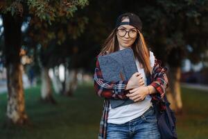 contento giovane femmina alunno vestito nel casuale capi di abbigliamento con tazza di caffè e zaino dietro a sua indietro a piedi in giro città. donna alunno Tenere il computer portatile e potabile caffè. estate tramonto volta. foto