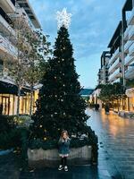 poco sorridente ragazza sta vicino un' Natale albero decorato con luminoso stelle su superiore foto