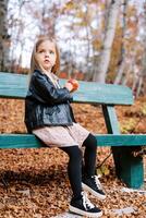 poco ragazza con un' rosso Mela nel sua mani si siede su un' di legno panchina nel il autunno foresta e sembra lontano foto