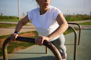 ritagliata Visualizza giovane sportivo donna esercizio all'aperto, fare braccia esercizi, spingendo su su attraversare bar nel urbano campo sportivo foto