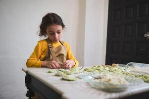 affascinante bambino ragazza fabbricazione Ravioli nel il casa cucina, vestito nel giallo maglione e beige dello chef grembiule foto