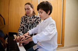 bello ispanico ragazzo nel bianca casuale camicia, apprendimento giocando pianoforte, seduta a di legno pianoforte forte e l'esecuzione un' musicale composizione sotto il guida di è insegnante. musicale formazione scolastica foto