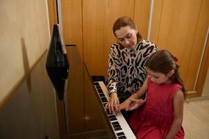 elementare età poco pianista ragazza giochi pianoforte, comporre un' melodia sotto il guida di sua insegnante durante individuale musica lezione a casa. bambini. formazione scolastica. stili di vita. Hobby e divertimento foto