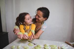 sorridente madre e figlia guardare a ogni Altro, Tenere scolpito fatti in casa Ravioli o ucraino varenyky, in piedi insieme a infarinato cucina tavolo, contro bianca parete sfondo foto