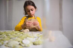 autentico bello poco casalinga, adorabile bambino ragazza nel dello chef grembiule, cucinando ravioli Ravioli con schiacciato patate, in piedi a cucina tavolo con ingredienti nel rustico accogliente domestico cucina foto