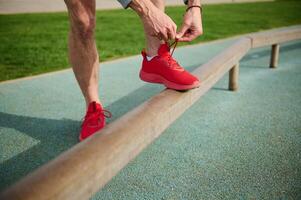 ritagliata Visualizza di un' in forma uomo legatura lacci delle scarpe di il suo rosso sportivo scarpe da ginnastica prima formazione o in esecuzione su il tapis roulant. foto