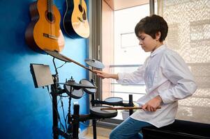 adolescenziale ragazzo nel bianca camicia e blu jeans, giocando tamburo kit nel il suo moderno musica studio camera. Due acustico elettrico chitarre sospeso su un' blu parete. le persone. musica. bambini Hobby e tempo libero attività foto