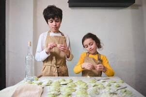 Due carino bambini fabbricazione Ravioli nel il casa cucina, vestito nel beige dello chef grembiuli. le persone. culinario. bambino apprendimento cucinando. infanzia. domestico vita foto