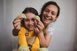 avvicinamento ritratto di un' madre e figlia avendo divertimento, sorridente guardare a telecamera, Tenere modellato Ravioli nel il casa cucina foto