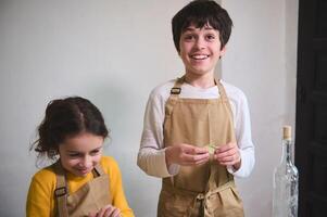 autentico ritratto di Due adorabile sorridente bambini fabbricazione fatti in casa Ravioli a casa cucina, in piedi contro bianca parete sfondo foto