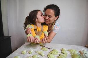 mamma e figlia avere divertimento, cucinando gnocco insieme. poco ragazza baci sua madre, contento per imparare il tradizionale ucraino culinario, mostrando modellato varennyky per telecamera, in piedi a infarinato tavolo foto