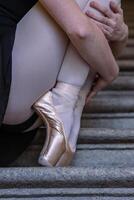 avvicinamento di ballerina indossare pointes scarpe Tenere sua mani su sua caviglie mentre seduta su il grigio pietra passi. danza riposo su il scala. ballerini piedi con rosa balletto pantofole Su. copia spazio foto