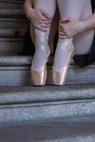 avvicinamento di ballerina pointes su il grigio pietra passaggi sfondo. danza seduta su il scala. ballerina è involucro sua mani in giro sua caviglie. ballerini piedi con rosa balletto pantofole Su. copia spazio foto