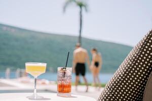 Due cocktail nel bicchieri in piedi su un' tavolo vicino un' sole lettino su il spiaggia contro il fondale di a piedi uomo e donna foto