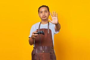 ritratto di un giovane asiatico che indossa un grembiule che tiene in mano il controller di gioco e fa l'arresto del movimento con le mani su sfondo giallo foto
