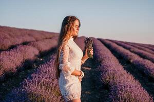 donna lavanda campo. contento spensierato donna nel un' bianca vestito a piedi nel un' lavanda campo e odorare un' lavanda mazzo su tramonto. ideale per caldo e ispirazione concetti nel voglia di girovagare e viaggio. foto